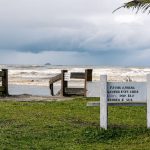Vista tranquila da praia de Peruíbe, com vegetação e uma placa de conservação sob céu nublado.