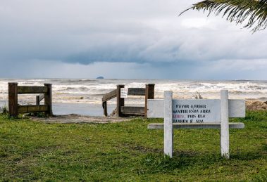 Vista tranquila da praia de Peruíbe, com vegetação e uma placa de conservação sob céu nublado.