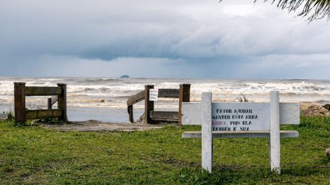 Vista tranquila da praia de Peruíbe, com vegetação e uma placa de conservação sob céu nublado.