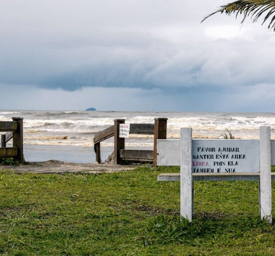 Vista tranquila da praia de Peruíbe, com vegetação e uma placa de conservação sob céu nublado.