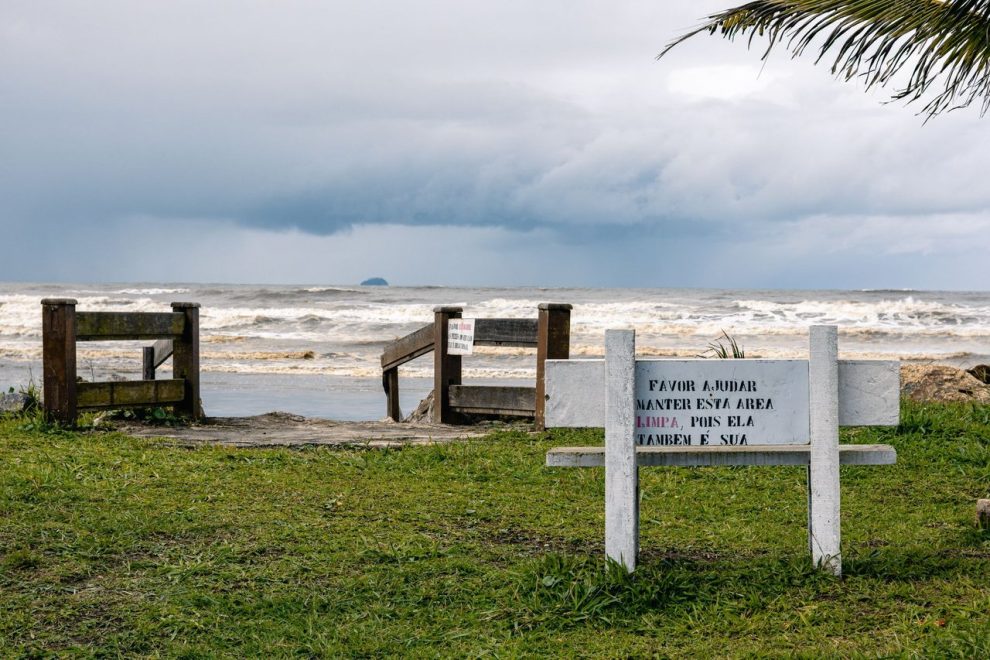Vista tranquila da praia de Peruíbe, com vegetação e uma placa de conservação sob céu nublado.
