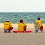 Três guarda-vidas uniformizados sentados na areia, voltados para o mar e observando a praia.