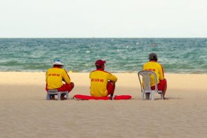 Três guarda-vidas uniformizados sentados na areia, voltados para o mar e observando a praia.