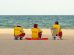 Três guarda-vidas uniformizados sentados na areia, voltados para o mar e observando a praia.