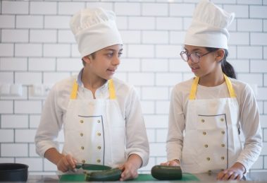 Duas crianças usando aventais e preparando vegetais em uma cozinha.