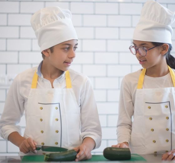 Duas crianças usando aventais e preparando vegetais em uma cozinha.