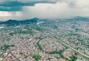 Vista aérea de uma extensa área urbana e de morros verdes sob a aproximação de uma tempestade.