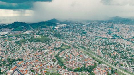 Vista aérea de uma extensa área urbana e de morros verdes sob a aproximação de uma tempestade.