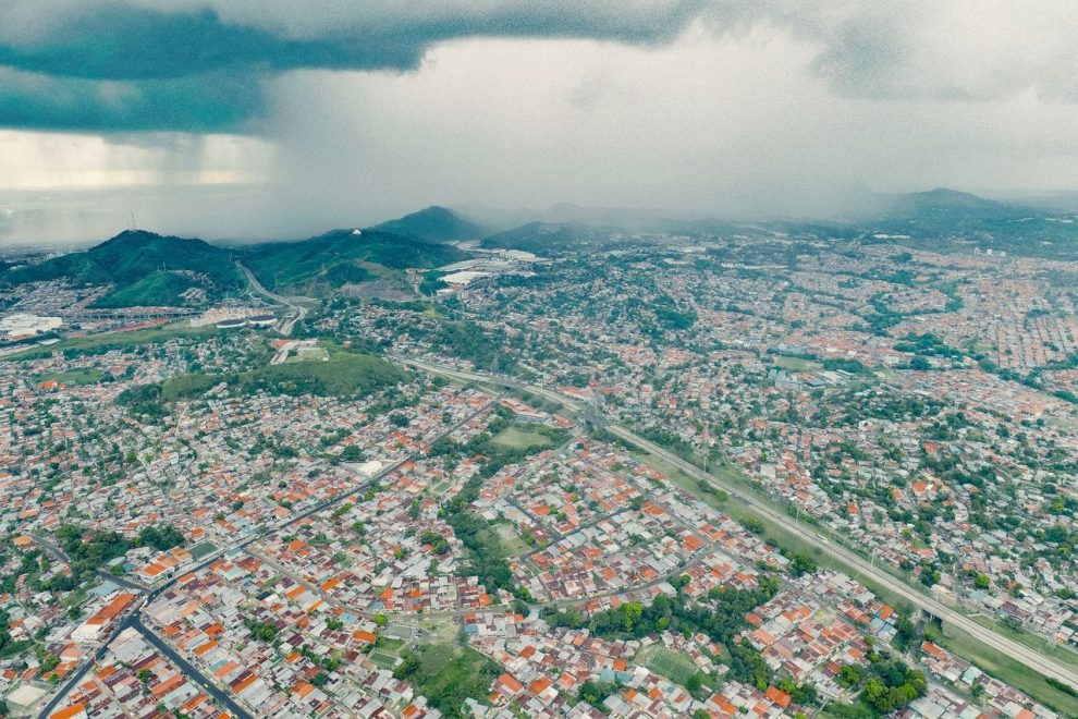 Vista aérea de uma extensa área urbana e de morros verdes sob a aproximação de uma tempestade.