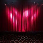 Palco de teatro vazio, com cortinas vermelhas e assentos ao fundo, preparado para uma apresentação.