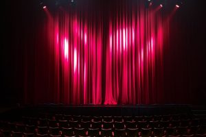 Palco de teatro vazio, com cortinas vermelhas e assentos ao fundo, preparado para uma apresentação.