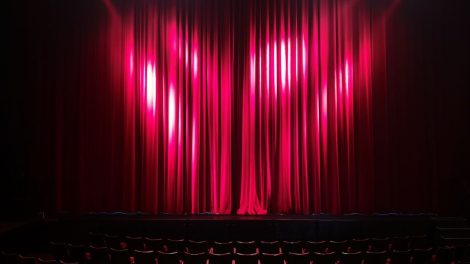 Palco de teatro vazio, com cortinas vermelhas e assentos ao fundo, preparado para uma apresentação.