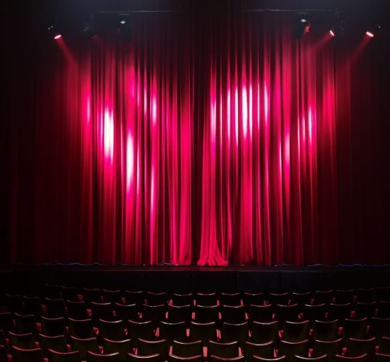 Palco de teatro vazio, com cortinas vermelhas e assentos ao fundo, preparado para uma apresentação.