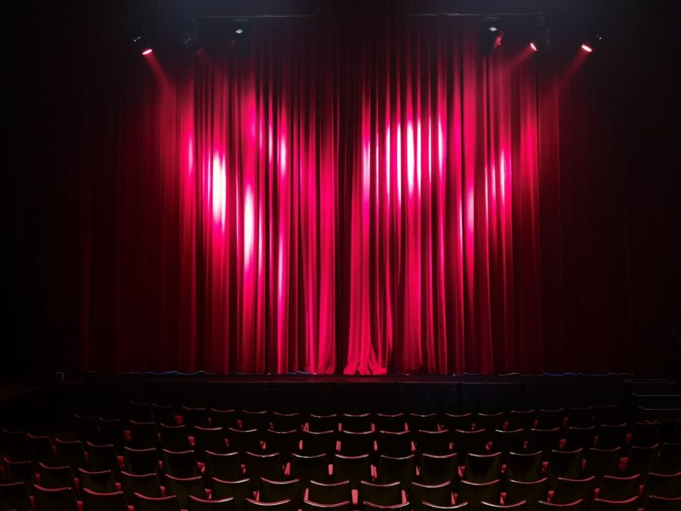 Palco de teatro vazio, com cortinas vermelhas e assentos ao fundo, preparado para uma apresentação.
