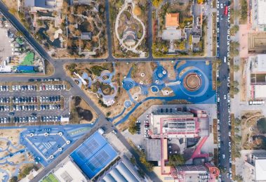 Vista aérea de um parque urbano com áreas de lazer, estacionamento e prédios ao redor