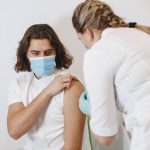 Profissional de saúde usando máscara aplica uma vacina no braço de um paciente em um consultório.