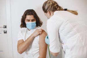 Profissional de saúde usando máscara aplica uma vacina no braço de um paciente em um consultório.