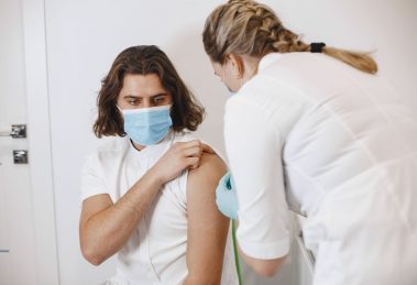 Profissional de saúde usando máscara aplica uma vacina no braço de um paciente em um consultório.