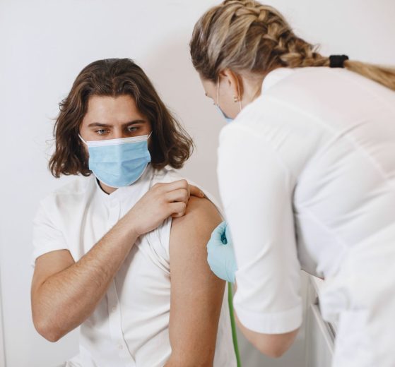 Profissional de saúde usando máscara aplica uma vacina no braço de um paciente em um consultório.