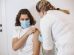 Profissional de saúde usando máscara aplica uma vacina no braço de um paciente em um consultório.
