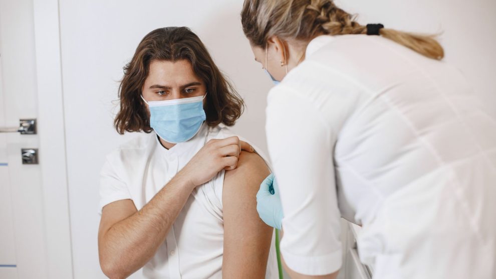 Profissional de saúde usando máscara aplica uma vacina no braço de um paciente em um consultório.
